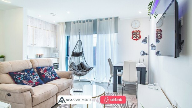 Un salon moderne avec un canapé, un fauteuil suspendu, un coin repas et une cuisine en arrière-plan.