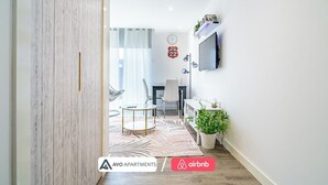 Un salon moderne avec un téléviseur, une table à manger et des chaises. On y trouve une plante sur le côté et une horloge au mur.