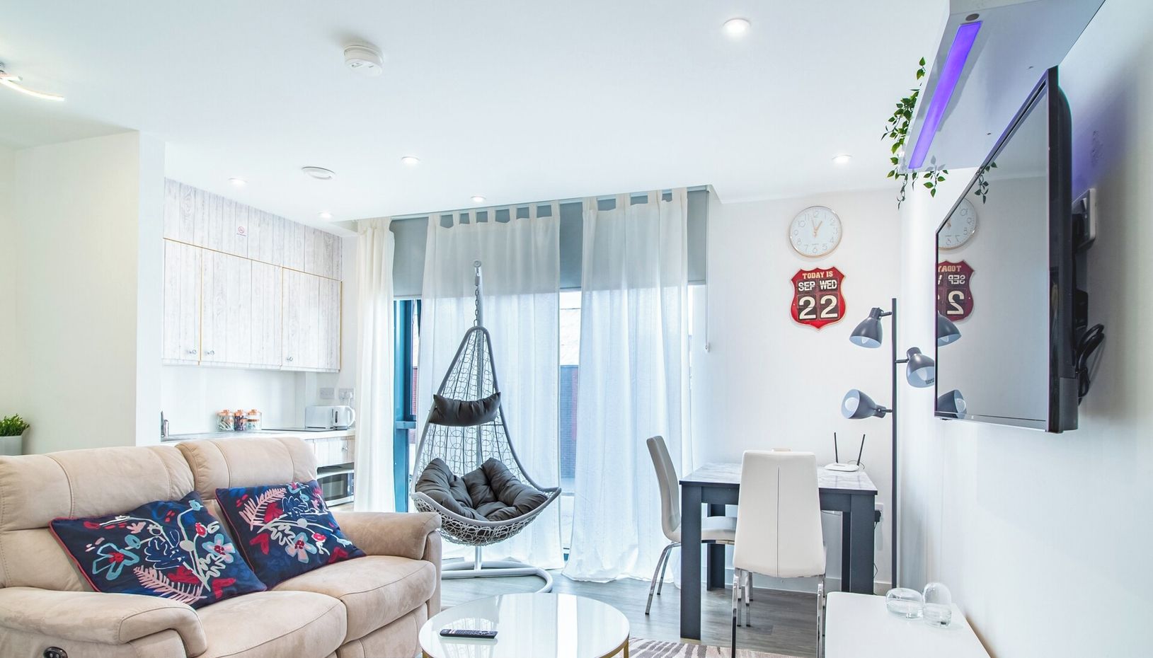 En moderne stue med sofa, hængestol, spiseplads og et køkken i baggrunden.