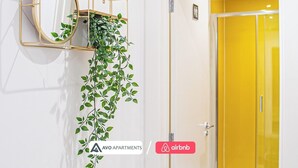 Een spiegel aan de muur, met een hangplant, een gele deur, en een bordje voor AVO APARTMENTS en Airbnb.