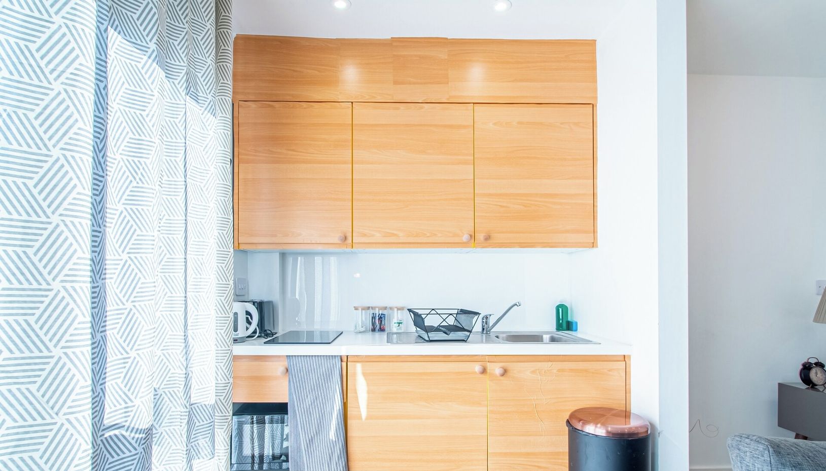 Eine moderne Küche mit Holzschränken, einer Arbeitsplatte und einem Spülbecken. Es steht ein Wasserkocher, eine Kaffeemaschine und ein Geschirrgestell bereit. Die Küche gehört zu einem AVO apartments Objekt, wie der Branding zeigt.