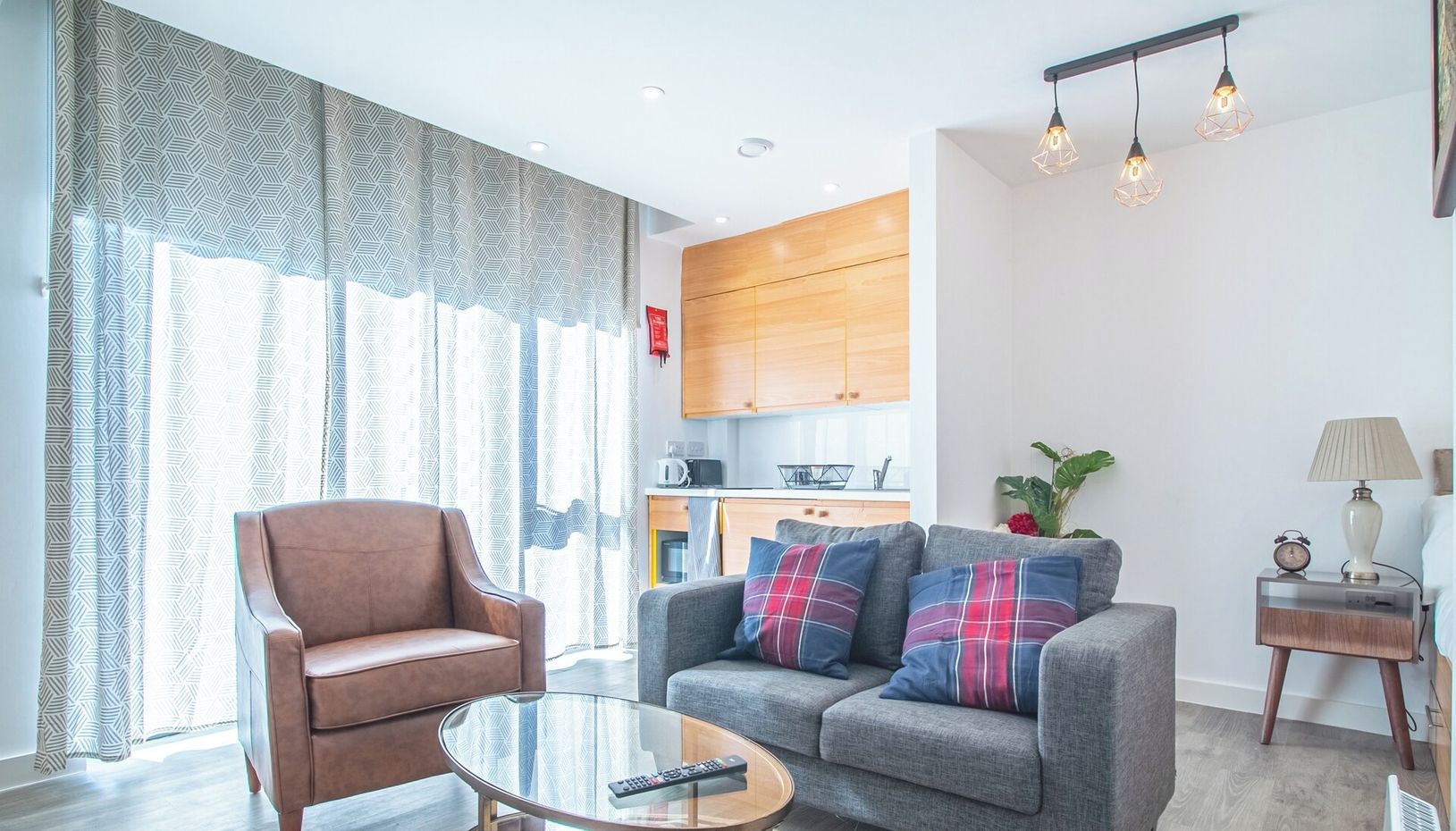 A modern living room with a brown leather sofa, a grey sofa with plaid cushions, a glass coffee table, and a kitchen in the background.