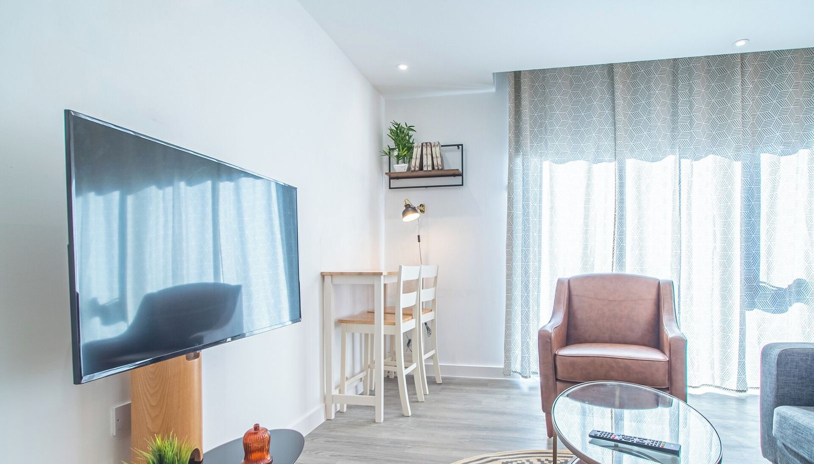 A modern living room with a flat-screen TV, a round coffee table, a sofa, and a chair. There is a small table with a plant and a lamp. The room has a large window with patterned curtains.