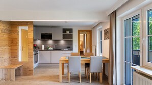 A modern kitchen with white cabinets, a dining area with a wooden table and chairs, and a large window with curtains.