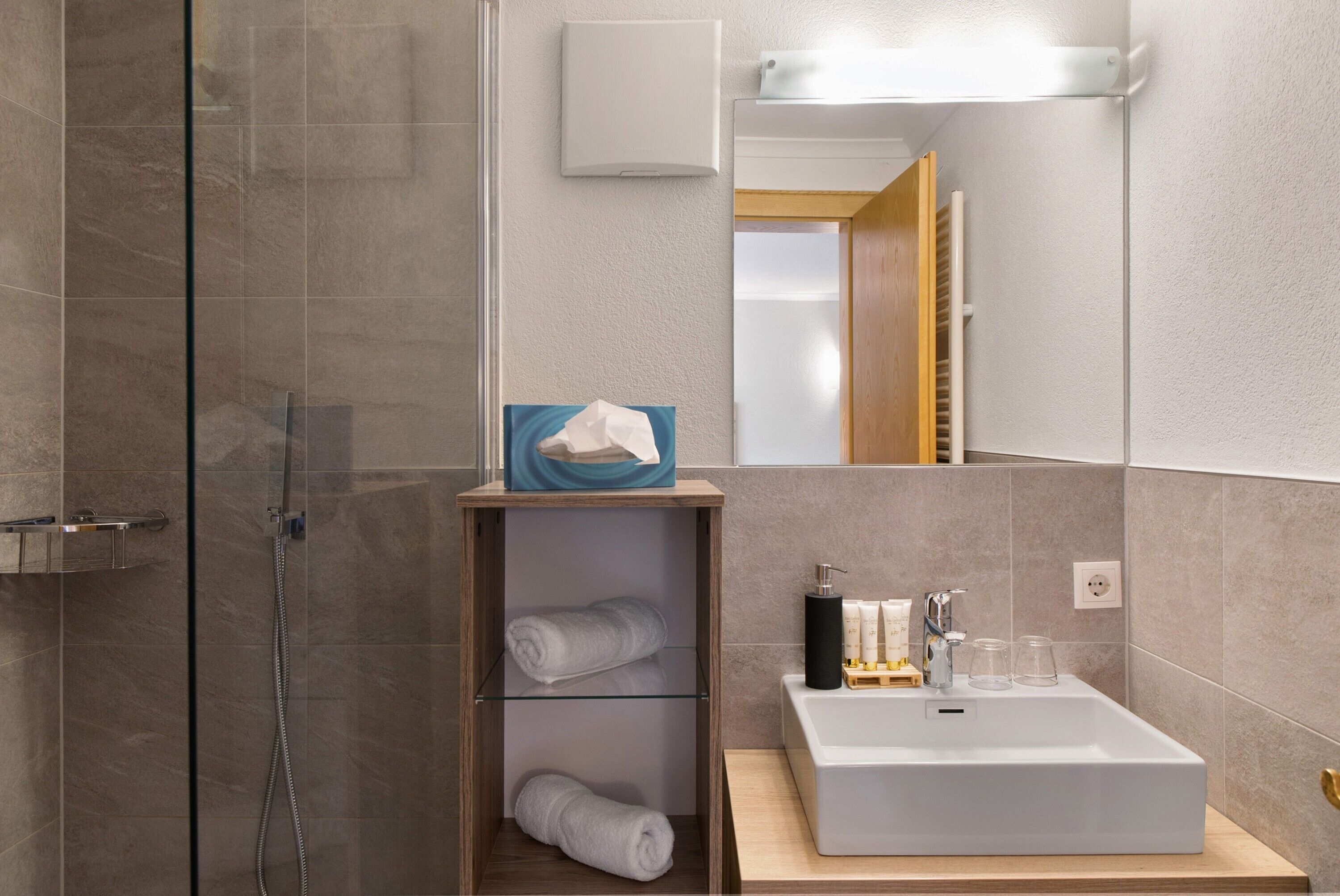 Une salle de bain moderne avec une douche à parois de verre, une étagère en bois où sont posées des serviettes, un lavabo blanc et un miroir.