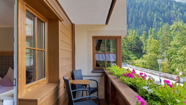 A balcony with a table and chairs, a window with a view of a forest, and a railing with flowers.