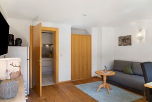 Een kamer met een bank, een klein rond tafeltje, een houten garderobe en een keukenruimte zichtbaar door een geopende deur.