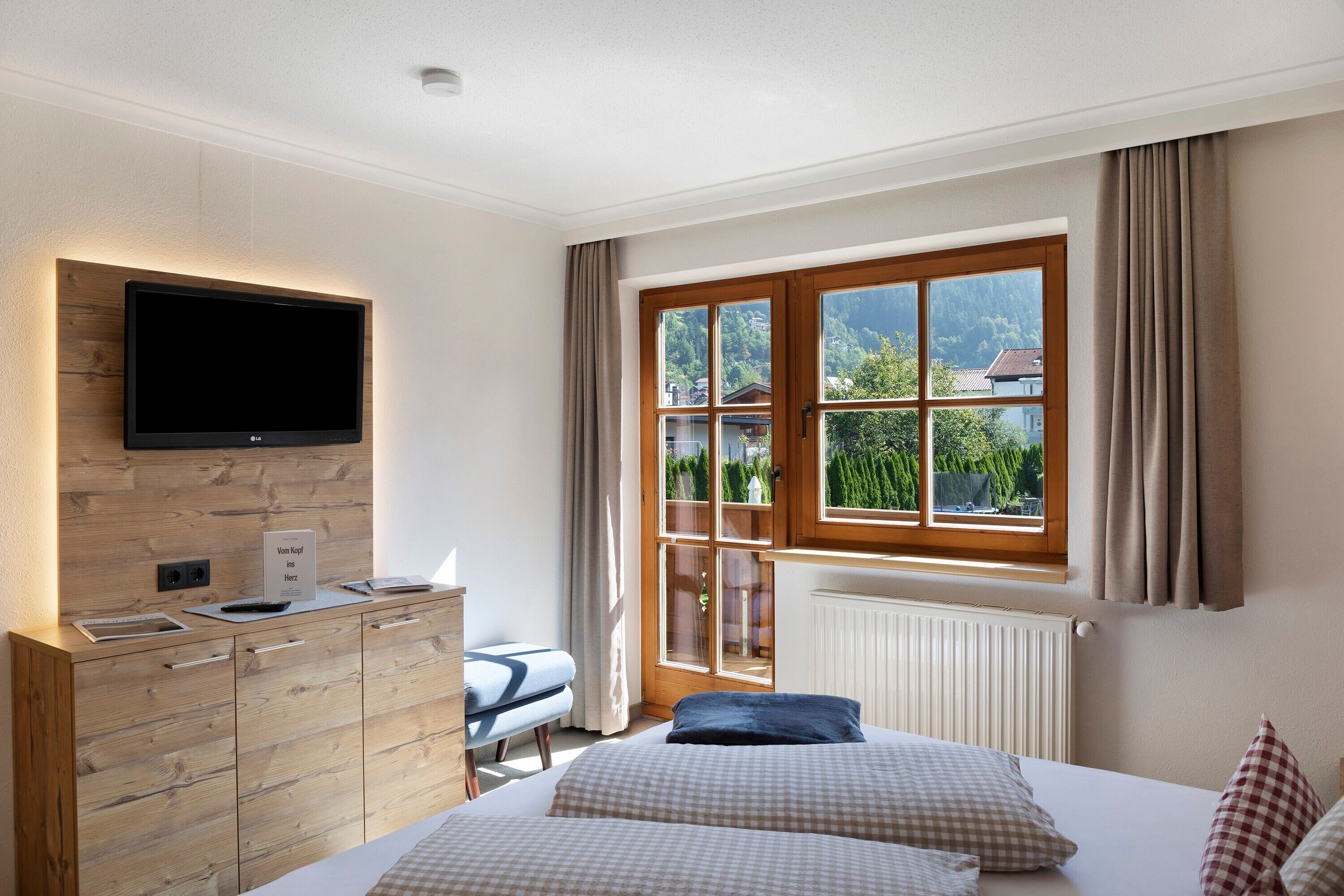 Une chambre avec un lit, un téléviseur fixé au mur en bois, une fenêtre avec des rideaux et une vue sur la verdure à l’extérieur.