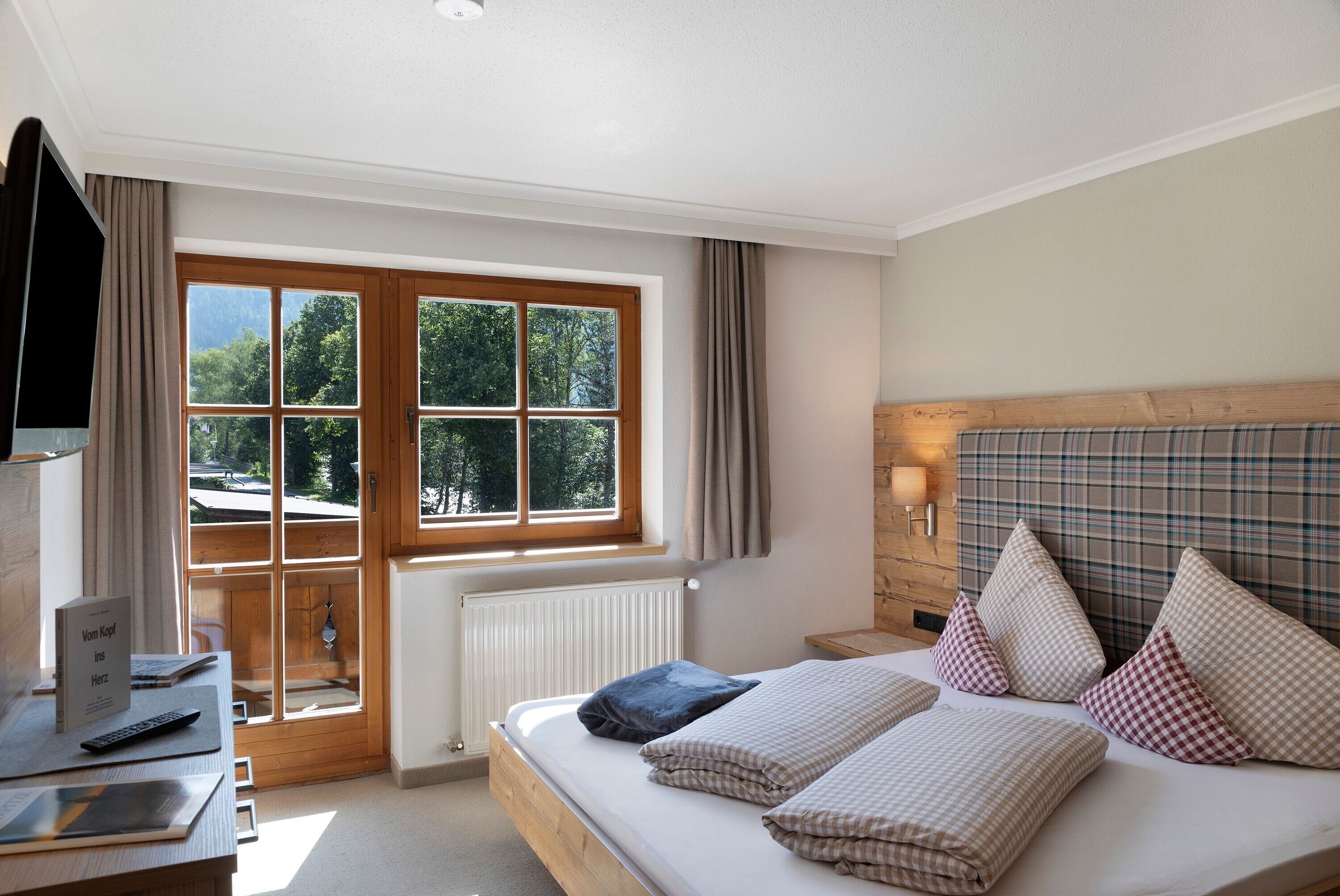 A bedroom with a wooden bed, a desk with a laptop, a window with curtains, and a view of trees.