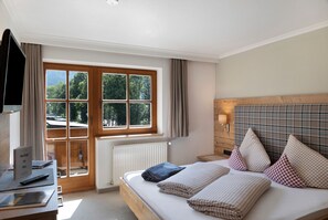 Een slaapkamer met een houten bed, een bureau met een laptop, een raam met gordijnen en uitzicht op bomen.