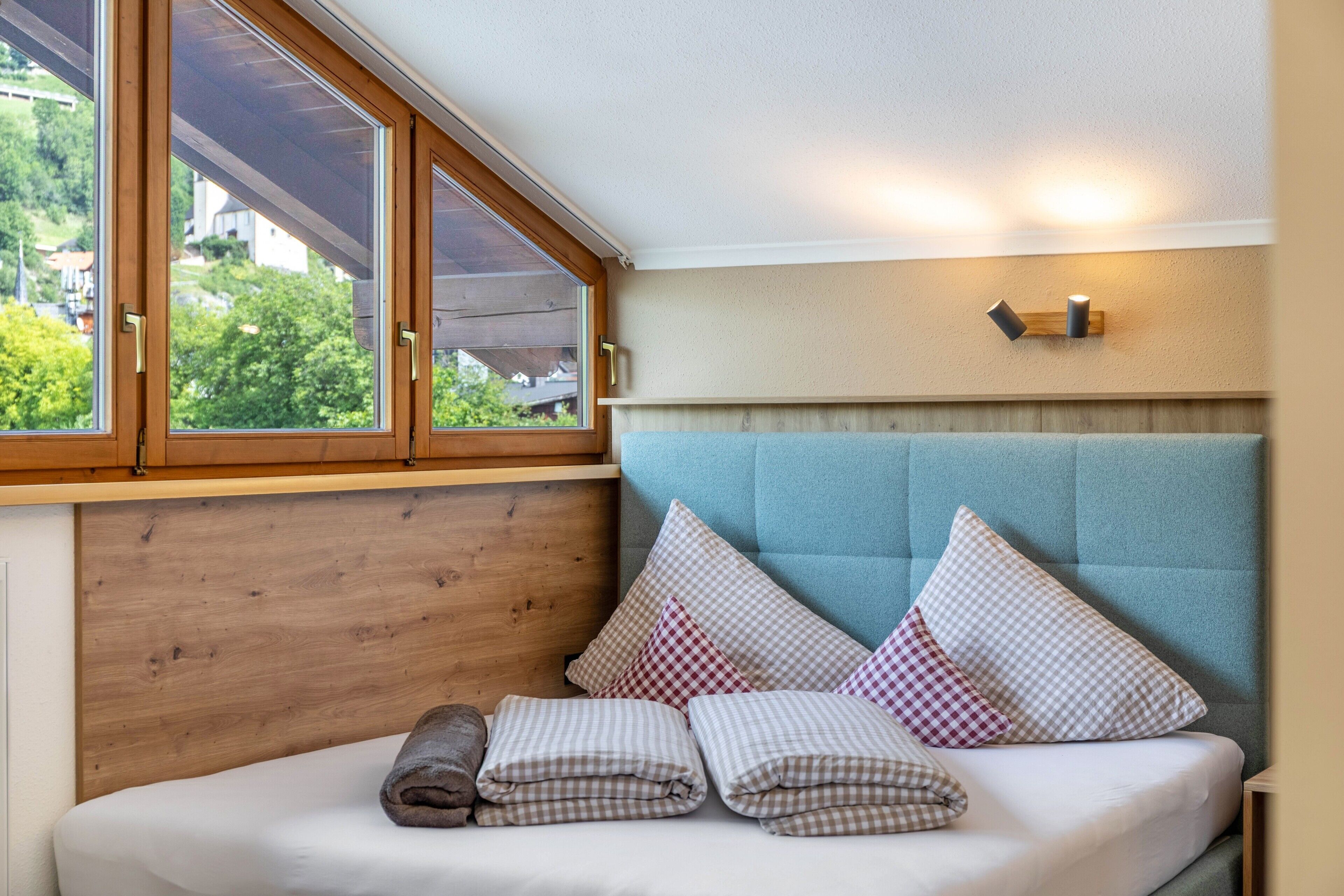 A bedroom with a wooden headboard, a window with blinds, a bed with checkered pillows, and a wall-mounted light fixture.