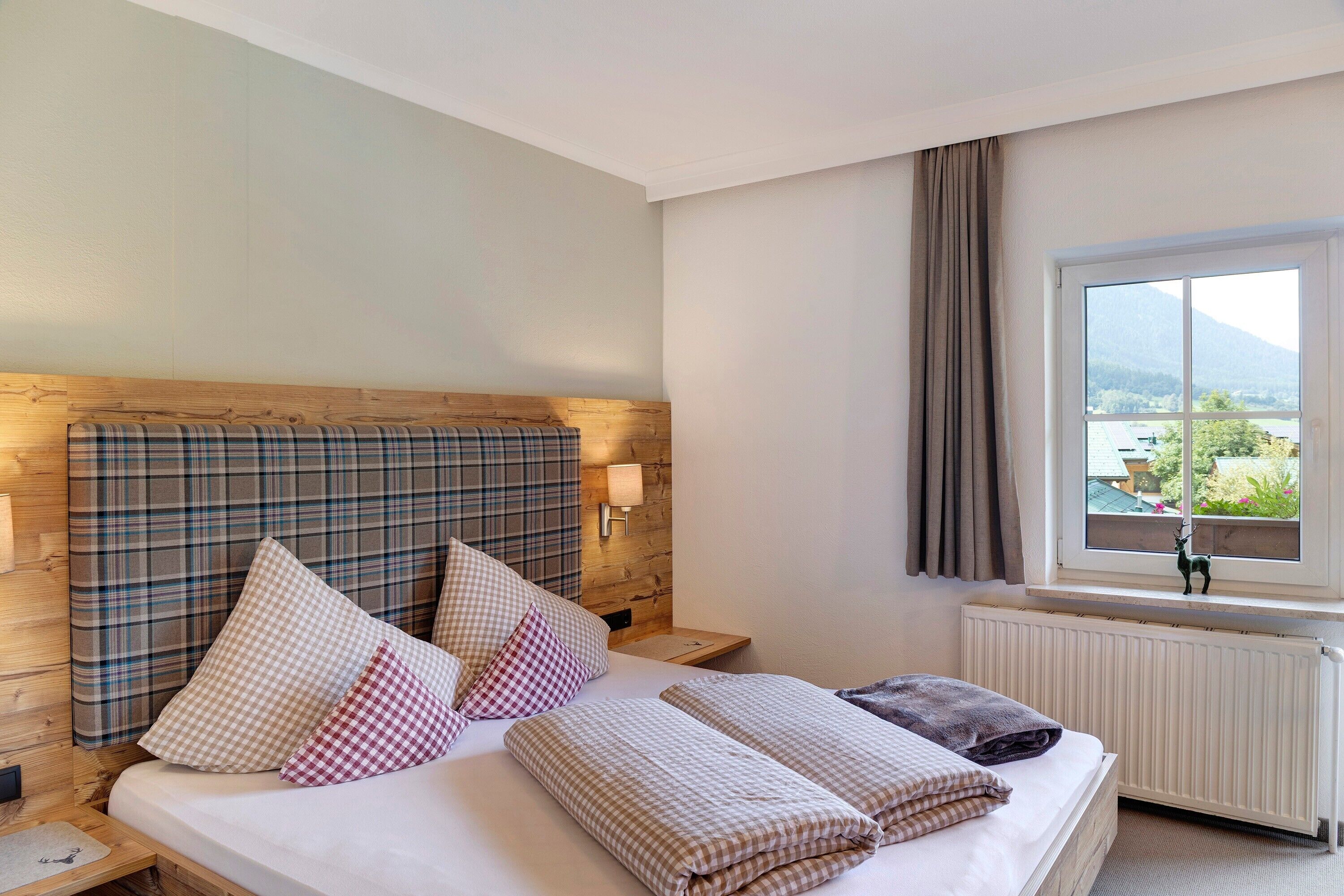 A hotel room with a bed, checkered pillows, a window with a view, a wooden headboard, and a nightstand.
