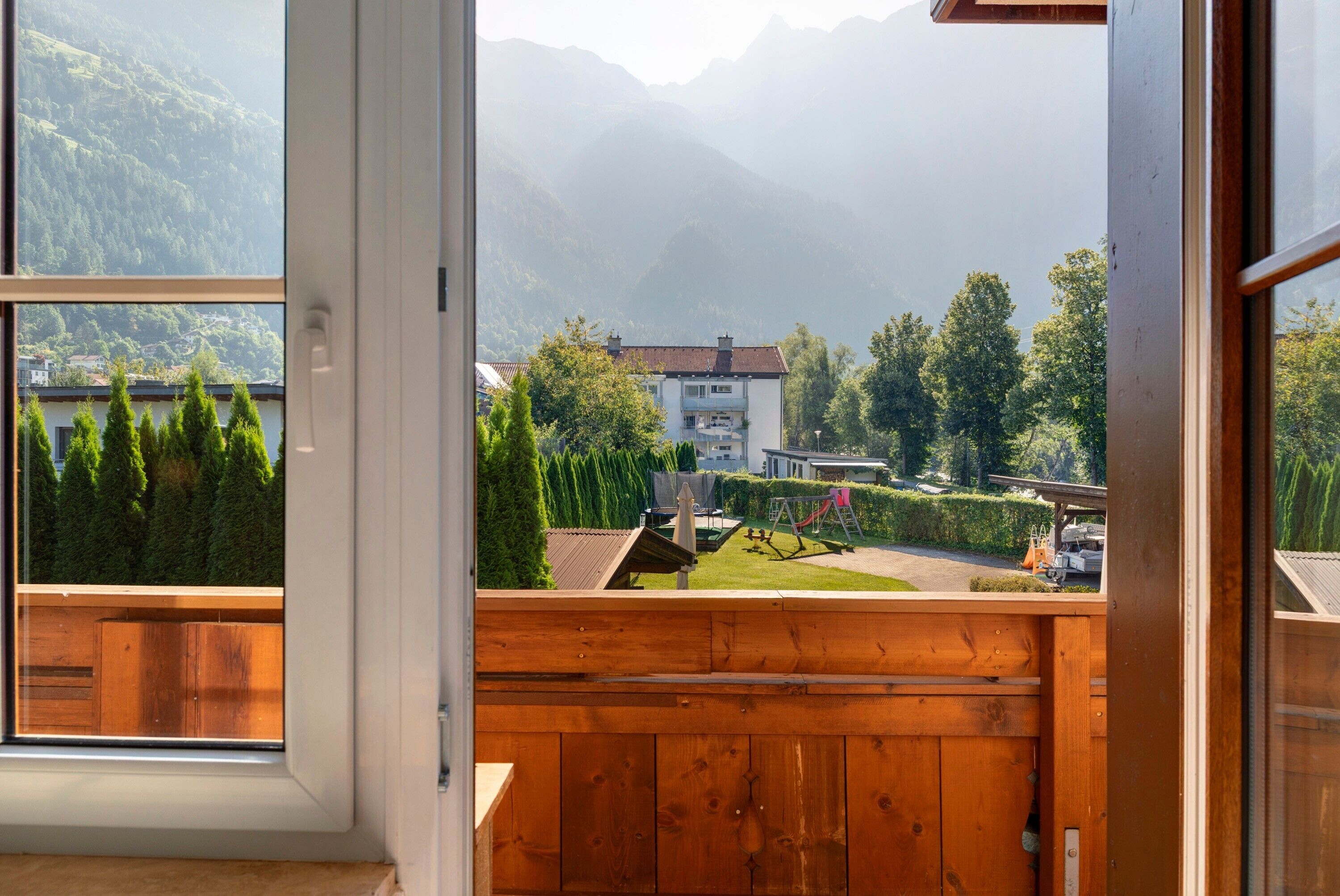 Une vue depuis un balcon doté d’une balustrade en bois, donnant sur un quartier résidentiel avec des maisons, des arbres et des montagnes en arrière-plan.