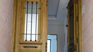 A wooden door with a window and a handle, leading into a hallway with a trash bin and a door in the background. - Yeşil Ege Otel (Ayvalik)