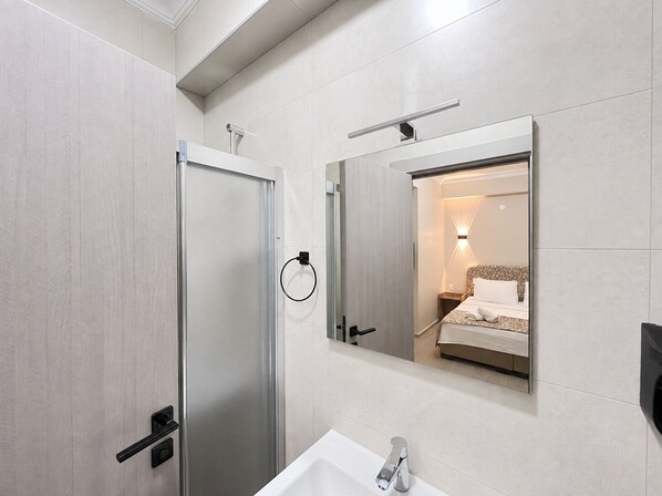 A modern bathroom with a shower, a sink, and a mirror reflecting a bed. - Yeşil Ege Otel (Ayvalik)