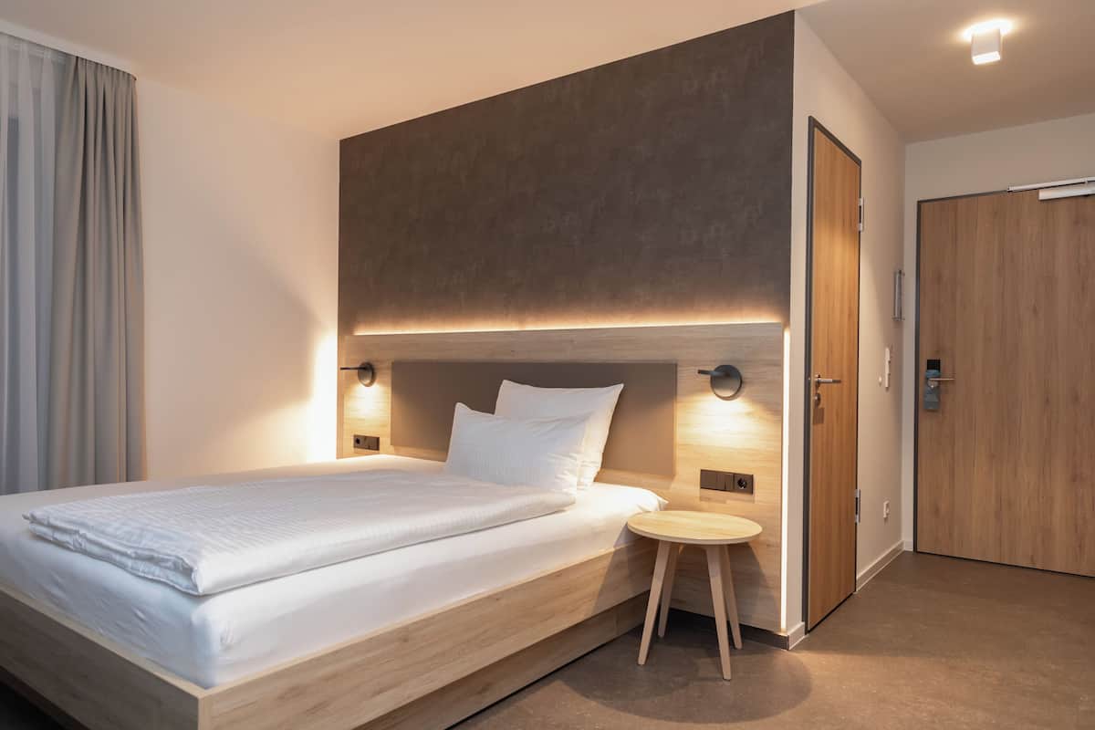 A modern hotel room with a large bed, bedside tables, a small stool, and a wooden wardrobe.