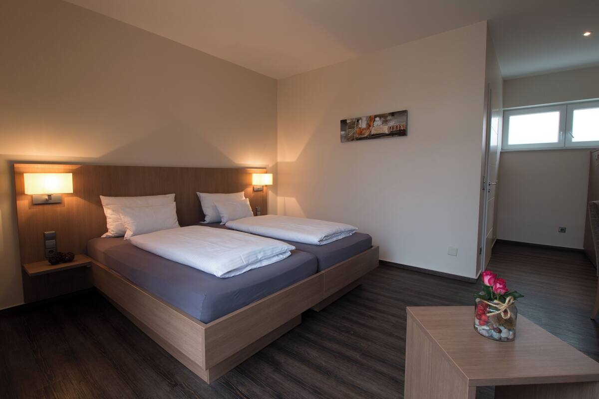 A modern hotel room with a double bed, bedside lamps, a small table with a vase, and a painting on the wall.