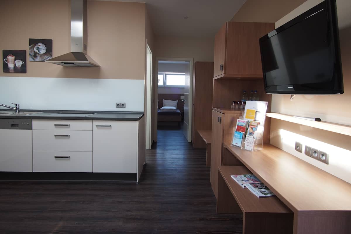 A modern kitchen with a stove, sink, and cabinets, a television mounted on a wooden shelf, and a hallway leading to a bedroom.