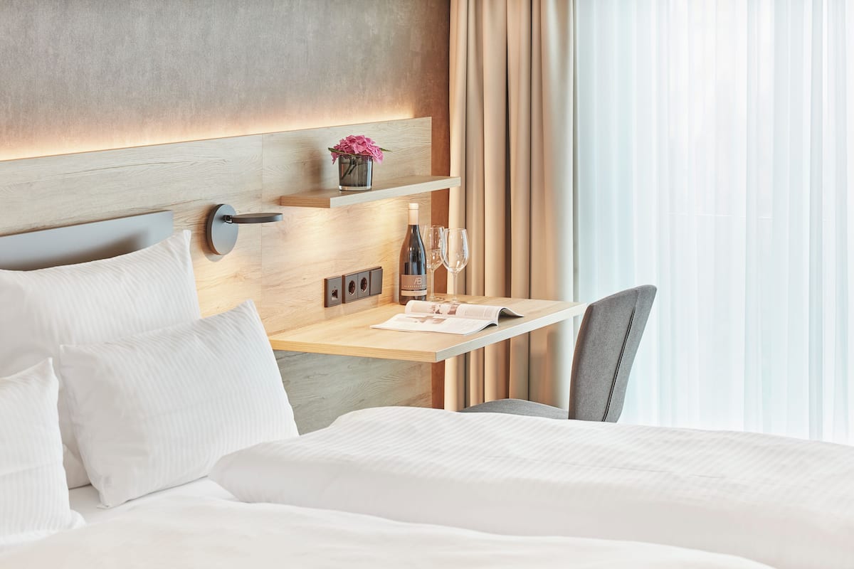 A hotel room with a bed, a bedside table with a bottle and glasses, a chair, and a wall-mounted shelf with flowers.