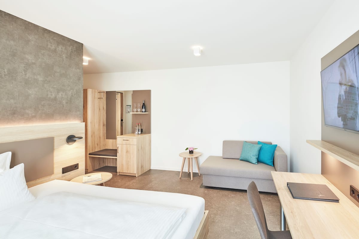 A modern hotel room with a bed, a desk, a sofa, a small table, a mirror, and a TV.