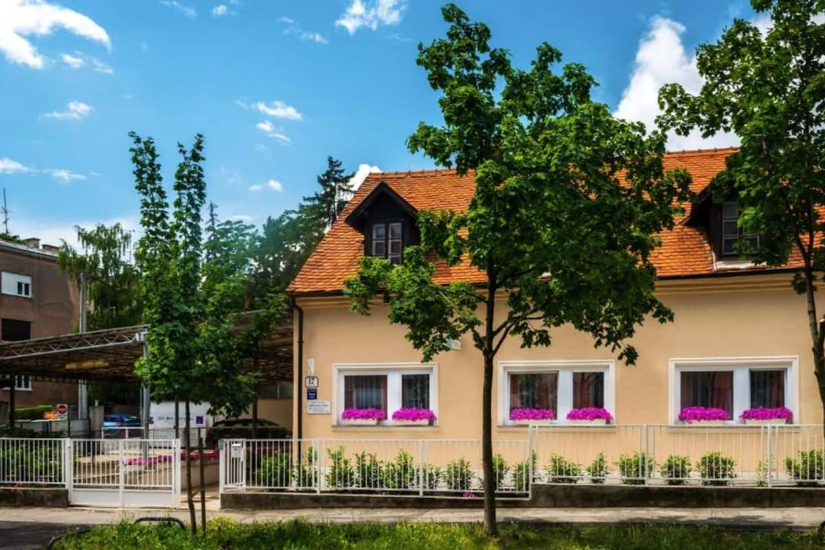 Ein zweistöckiges Gebäude mit rotem Ziegeldach, weißen Fensterrahmen und einer beige farbenen Fassade. Vor den Fenstern stehen drei Töpfe mit violetten Blumen.