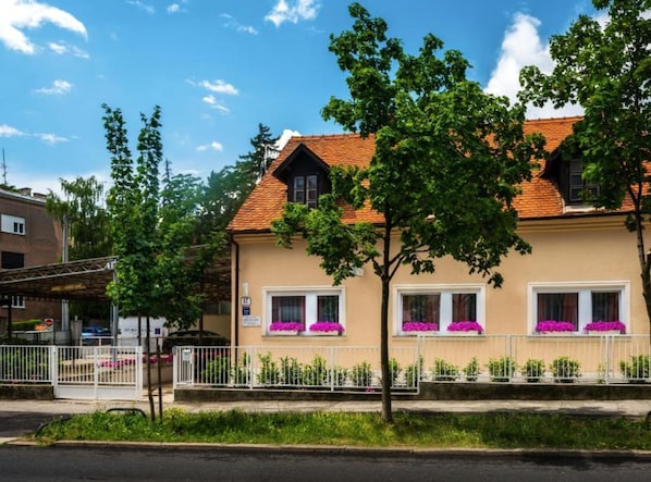 A two-story building with a red-tiled roof, white window frames, and a beige facade. There are three windows with purple flowers in front.