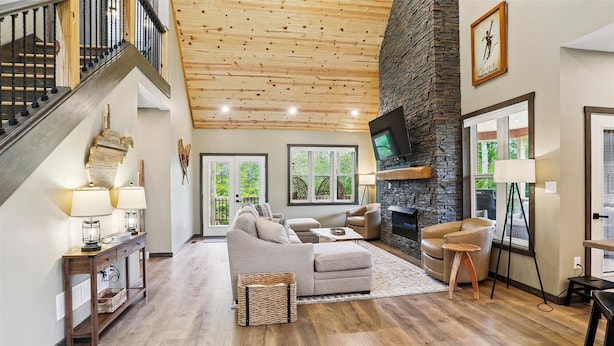 A spacious living room with a stone fireplace, a staircase, and large windows offering a view of the outdoors.