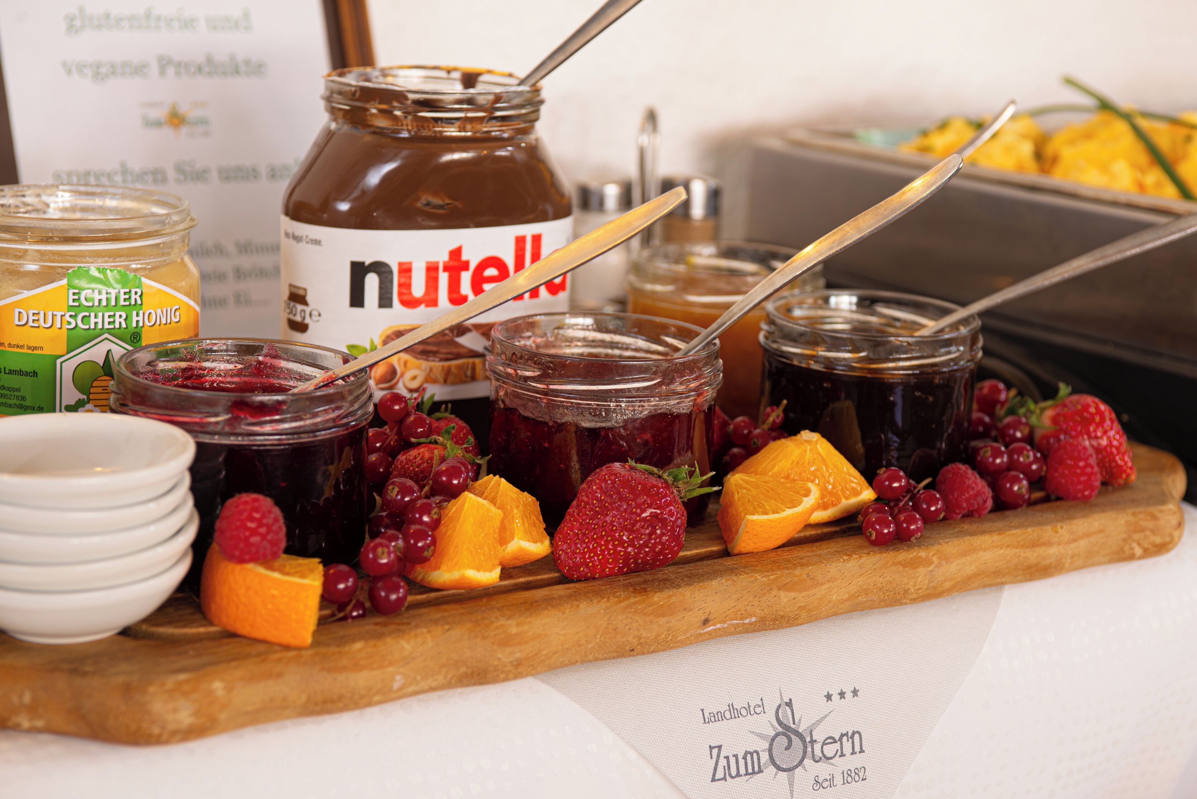 A variety of food items including jars of jam, a jar of Nutella, and fresh fruits like strawberries and grapes on a wooden board.