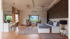 Quarto de hotel moderno com uma cama grande, uma escrivaninha, duas poltronas, um sofá e vista para o verde.