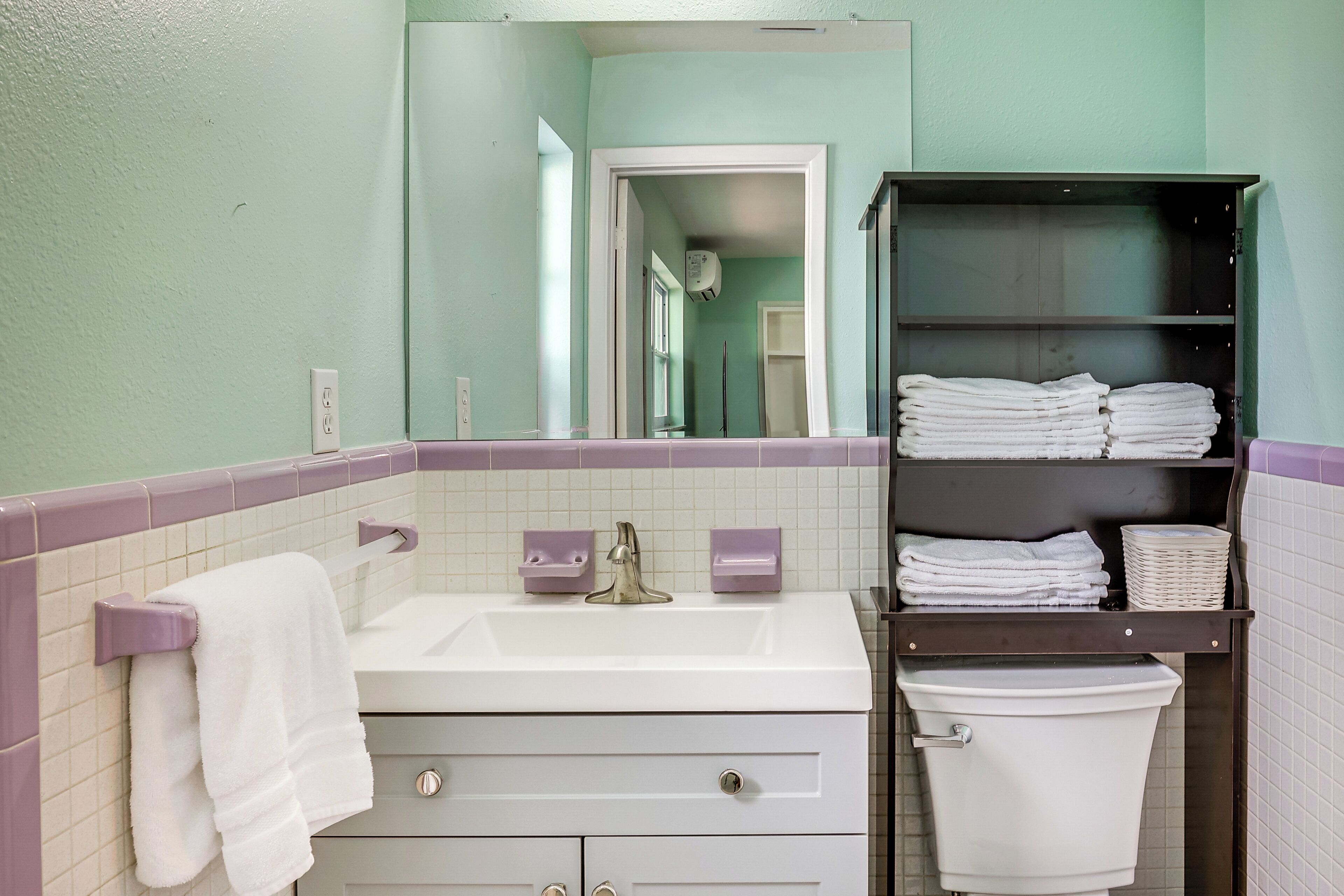 A bathroom with a white sink, a toilet, and a towel rack. There is a mirror above the sink and a shelf with folded towels.