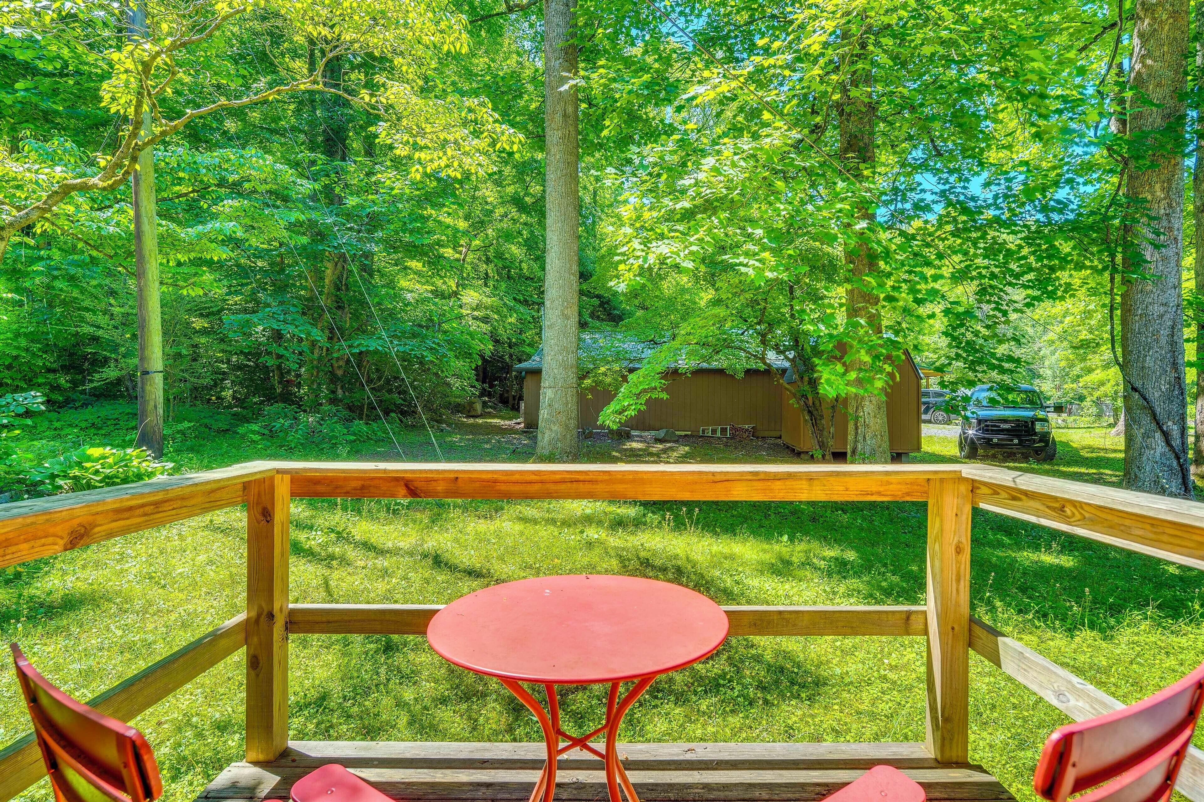 A wooden deck with a red table and chairs, surrounded by lush greenery and trees.