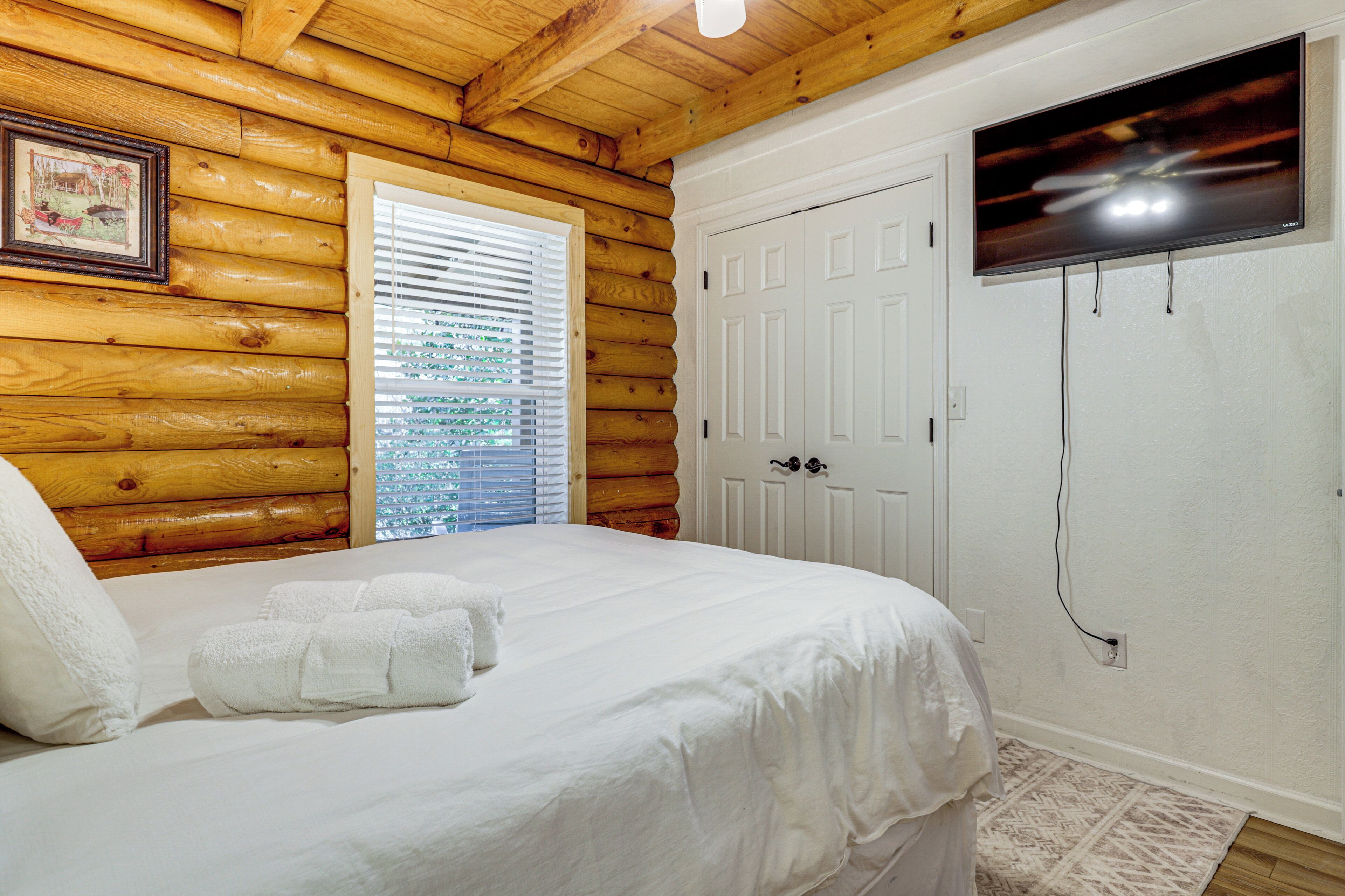 Un dormitorio de estilo cabaña de madera con una cama, ropa de cama blanca, un televisor montado en la pared y una ventana con persianas.