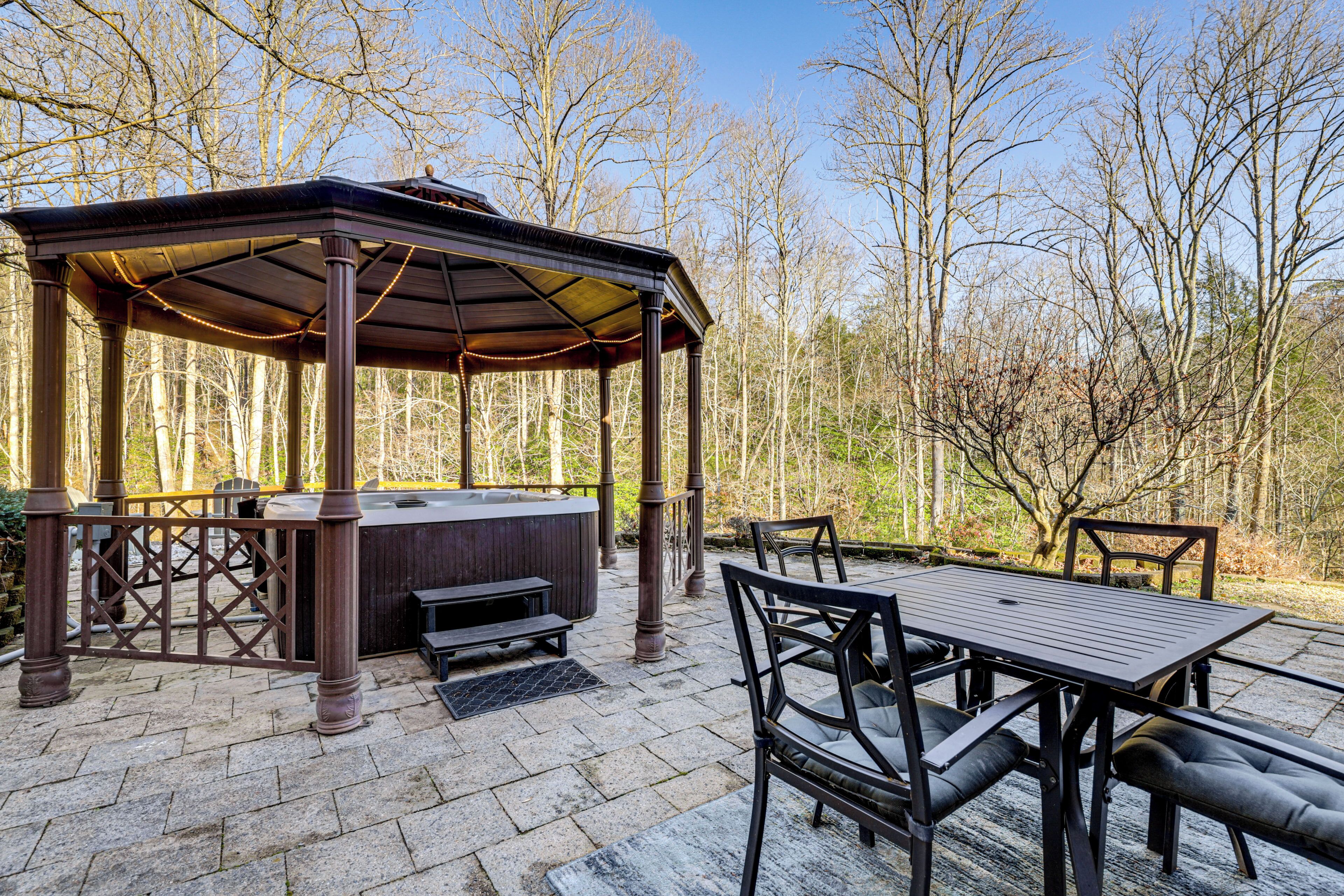 An outdoor patio with a wooden gazebo, a hot tub, and a dining table with chairs.
