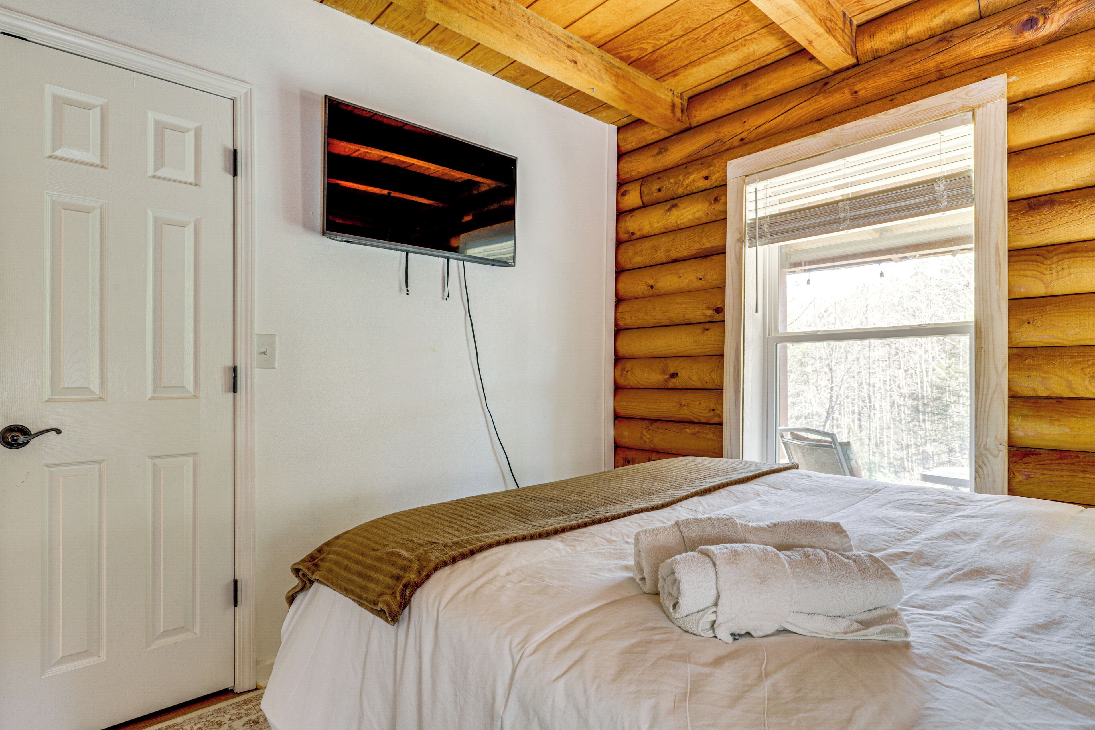 Une chambre avec un plafond en bois, une porte blanche, un téléviseur fixé au mur et une fenêtre avec des stores.