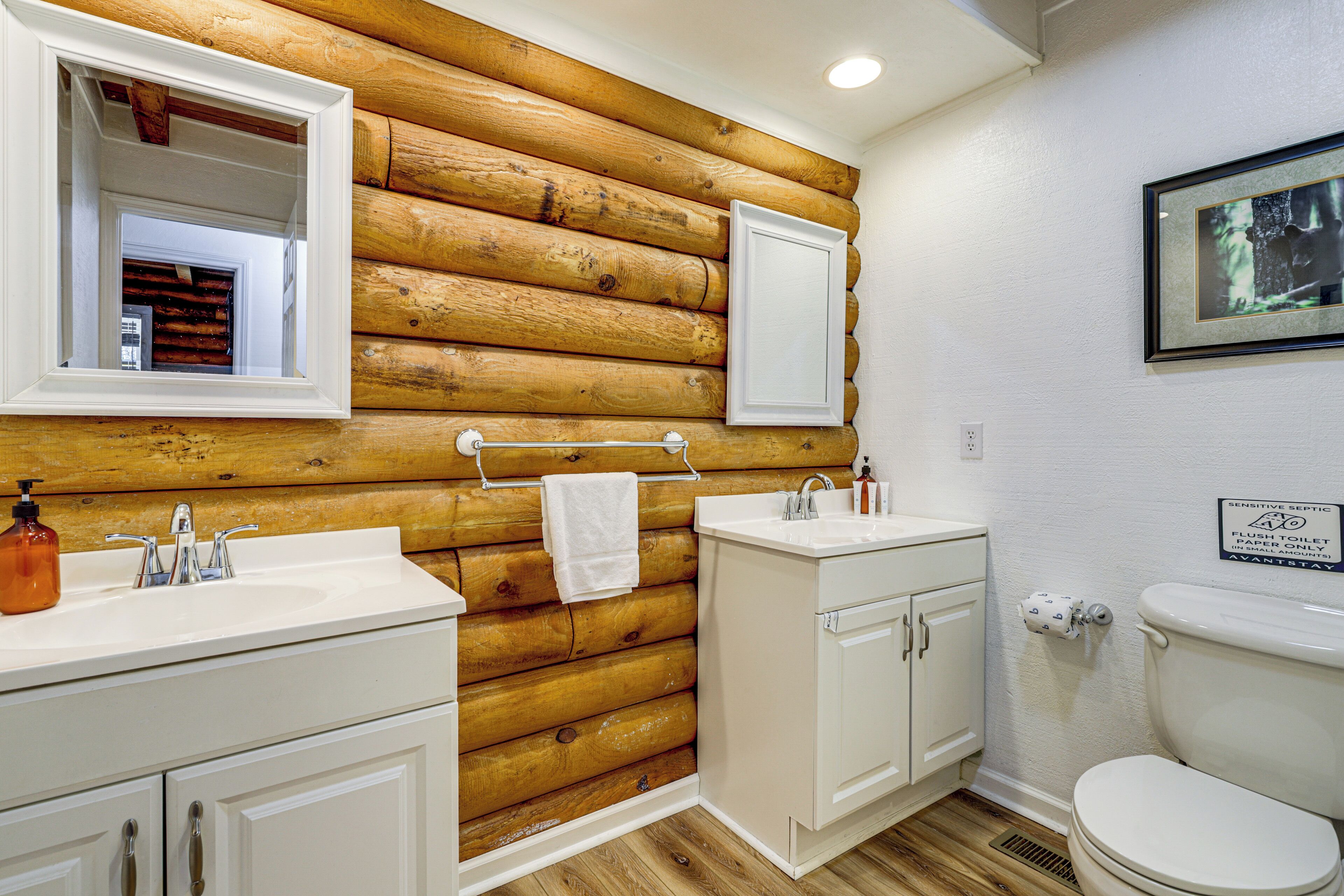 A bathroom with wooden log wall, white cabinets, a sink, and a toilet.