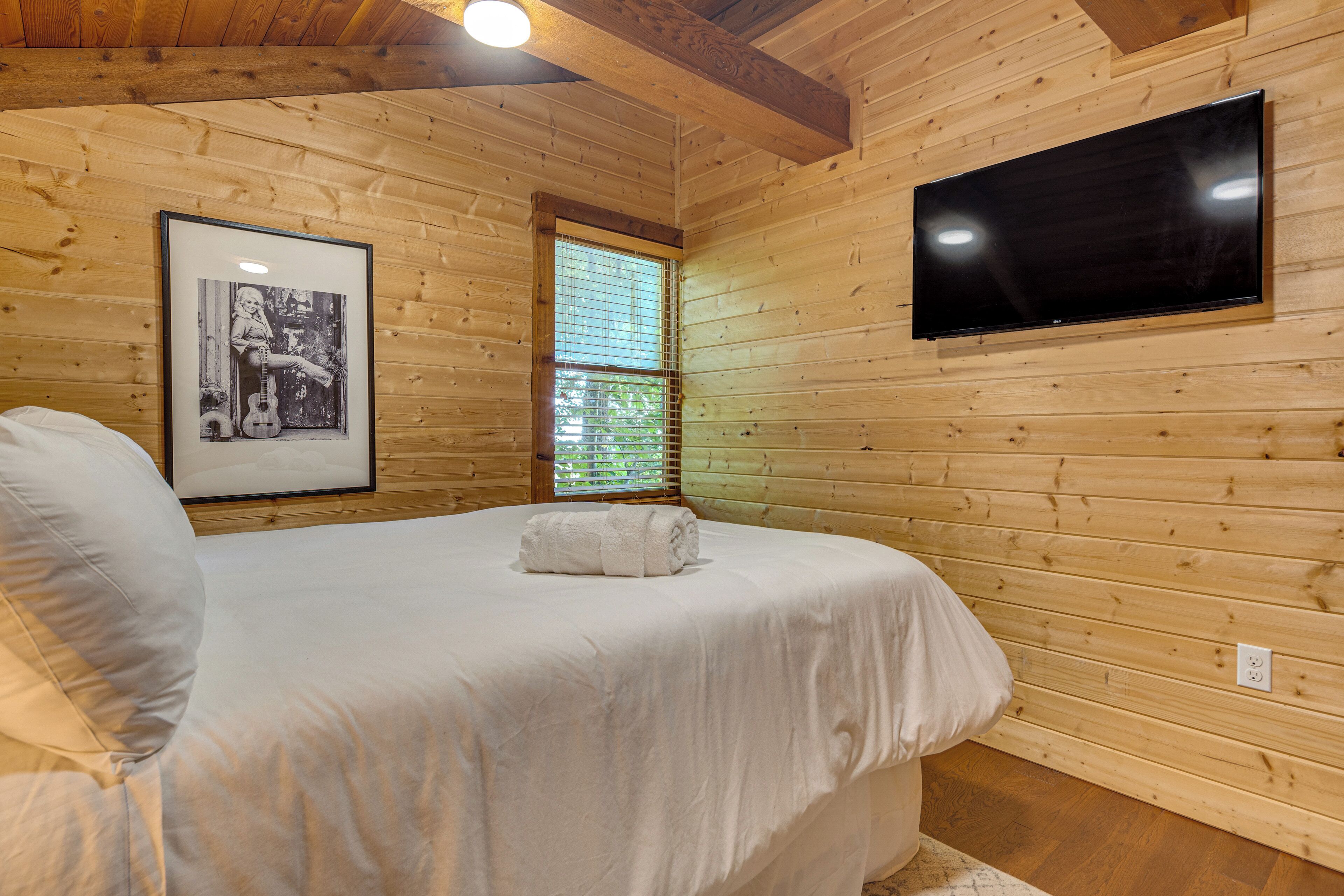 Een kamer met houten lambrisering, een bed, een ingelijst schilderij, een raam en een aan de muur bevestigde televisie.