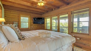 Une chambre dans une cabane en bois, avec un lit, un ventilateur de plafond, une télévision et de grandes fenêtres.