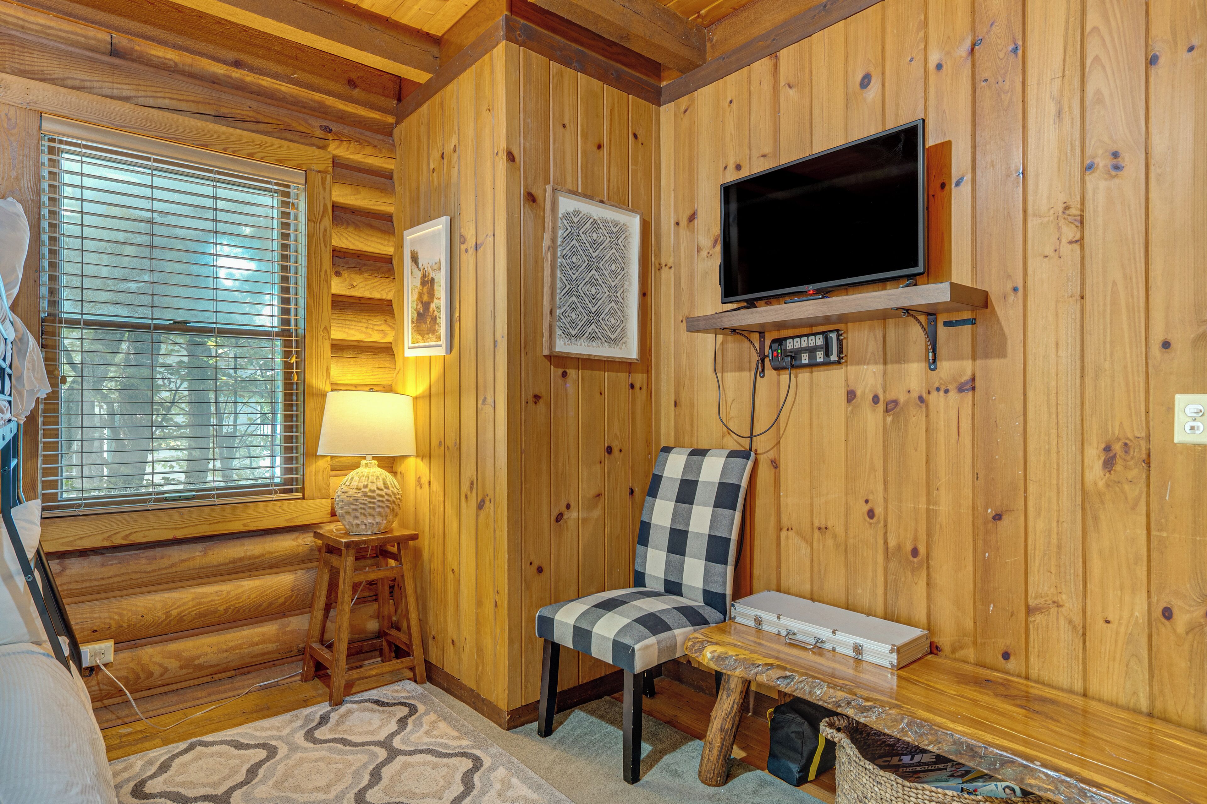 Ein Interieur einer Blockhütte mit einem an einer Holzablage montierten Fernseher, einem karierten Sessel, einem kleinen Tisch und einem Fenster mit Jalousien.
