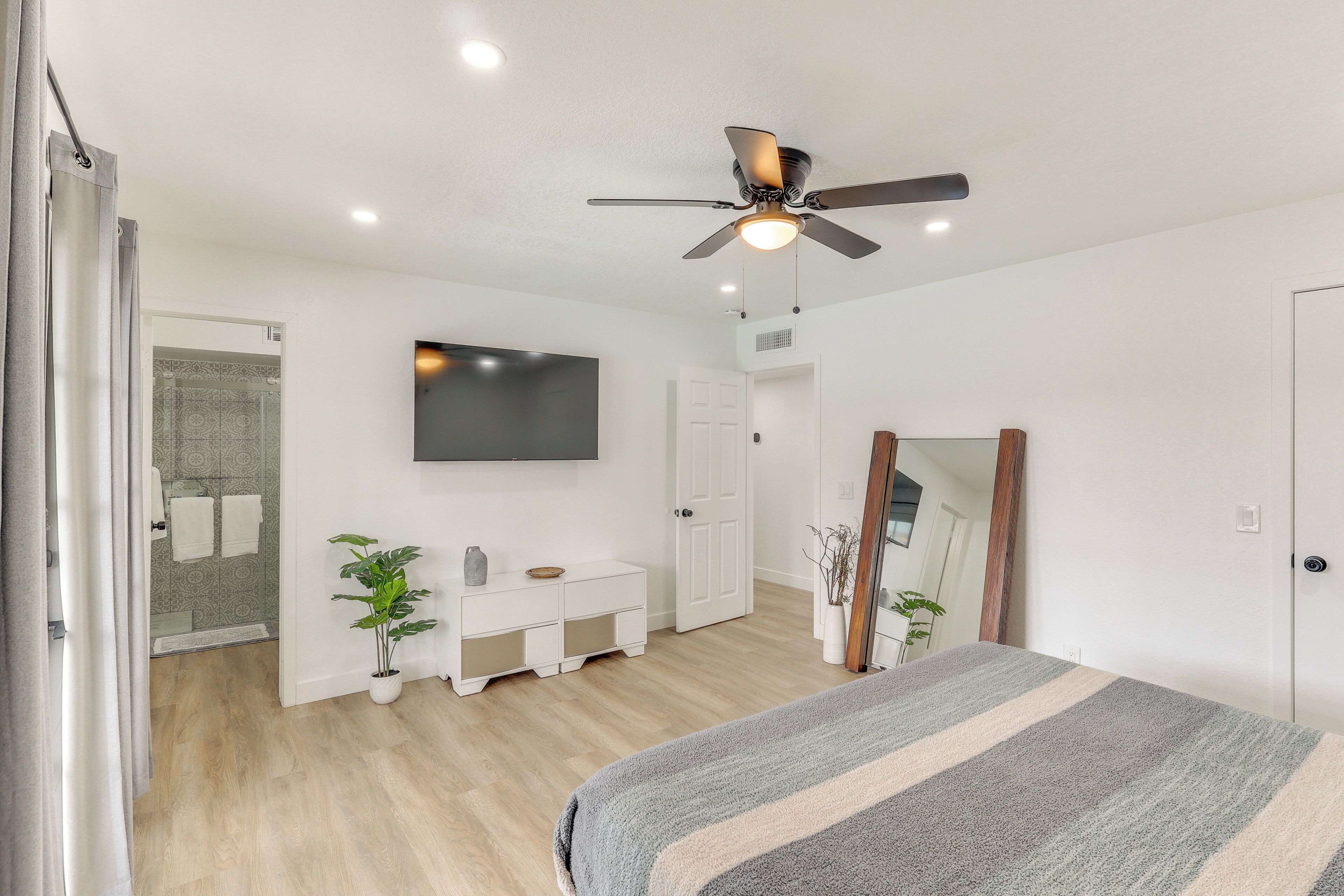 Un dormitorio con cama, ventilador de techo, televisor, armario blanco, mesita de consola y planta en maceta.