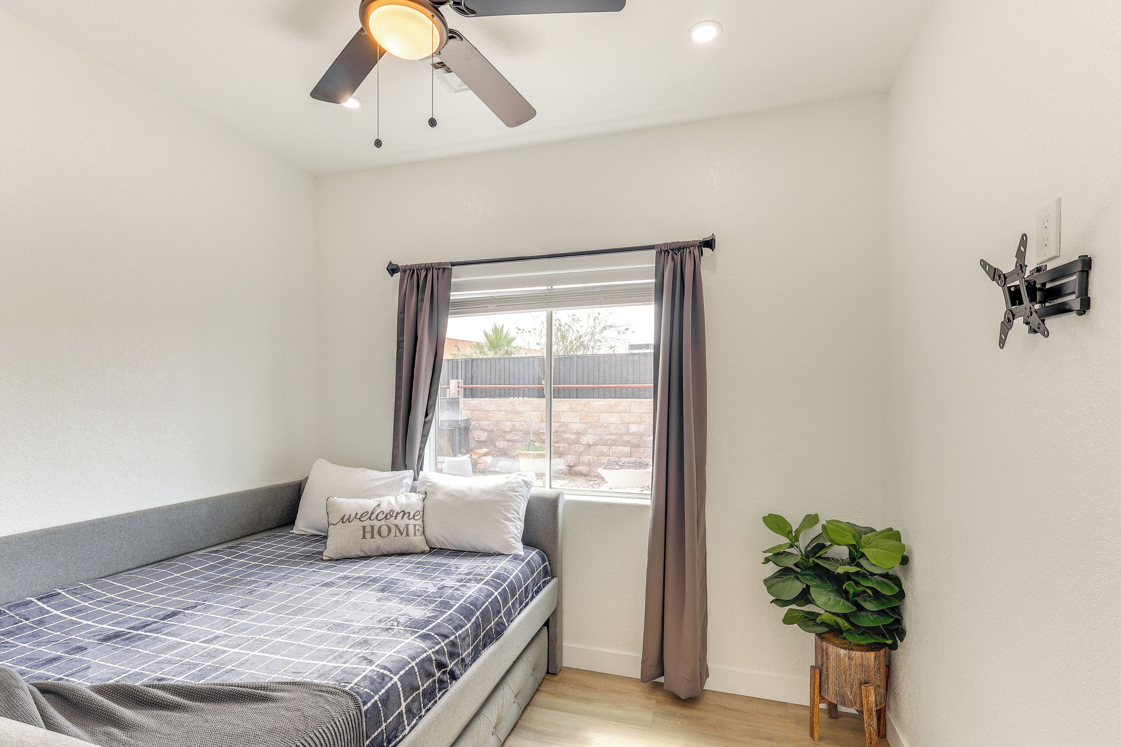 Une chambre à coucher avec un lit, un ventilateur de plafond, une fenêtre avec des rideaux, un petit tabouret en bois et une plante en pot.
