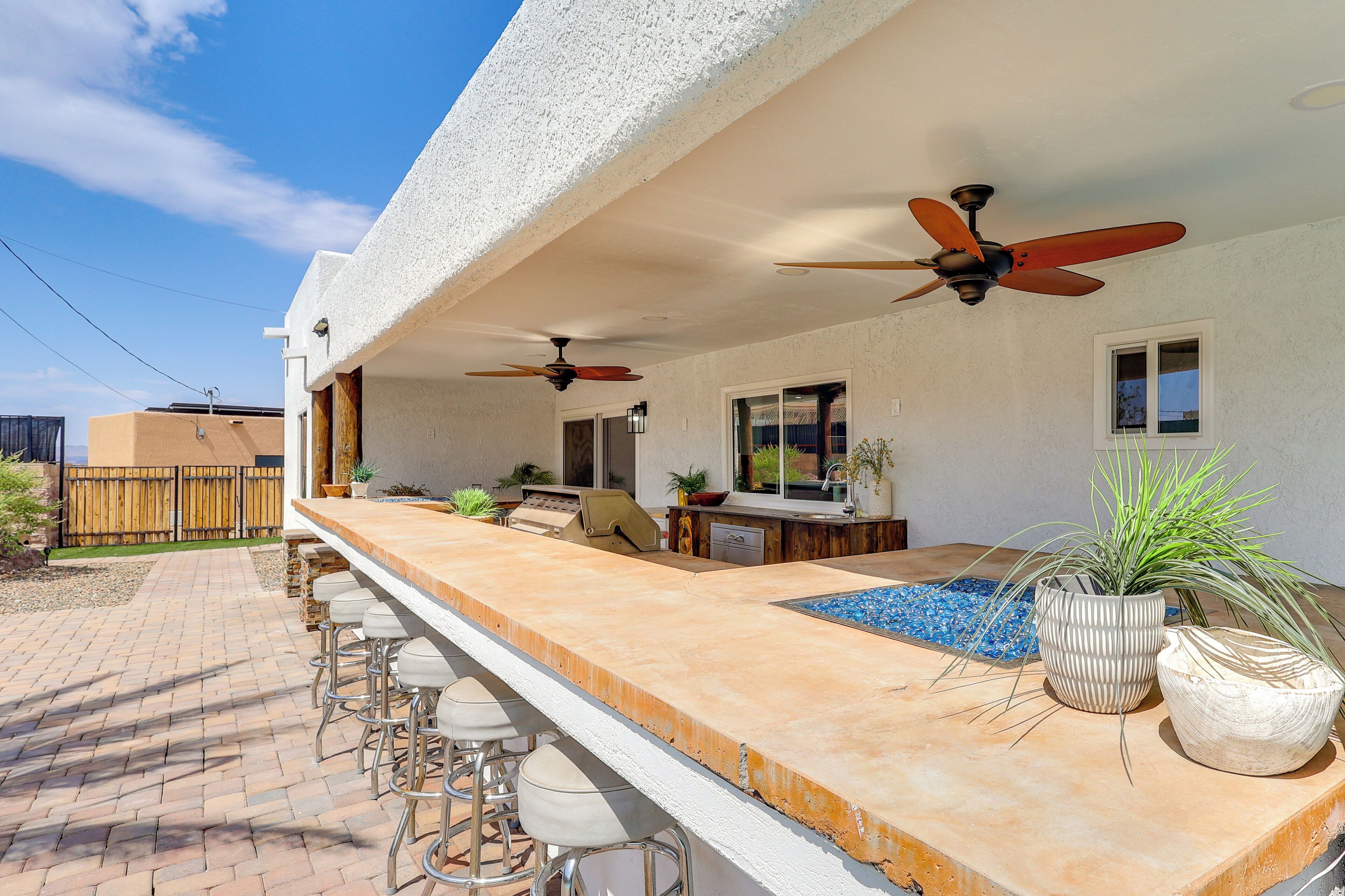 Un área de bar al aire libre con piscina, taburetes de bar, ventiladores de techo y plantas en macetas.