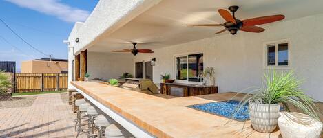 Uma área de bar ao ar livre, com piscina, banquetas, ventiladores de teto e plantas em vasos.