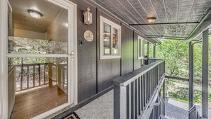 Un porche cubierto con barandilla de madera, vista a un bosque y una puerta que da a una habitación con piso de madera.