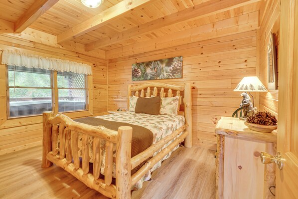 A wooden cabin bedroom with a bed, a nightstand, a lamp, and a painting on the wall. - Private Hot Tub: Lake Resort Cabin in Sevierville! (Sevierville)