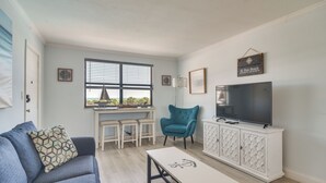 A living room with a sofa, a blue armchair, a TV, a cabinet, and a window with blinds. - Steps to St Pete Beach: Condo w/ Pool Access (St. Pete Beach)