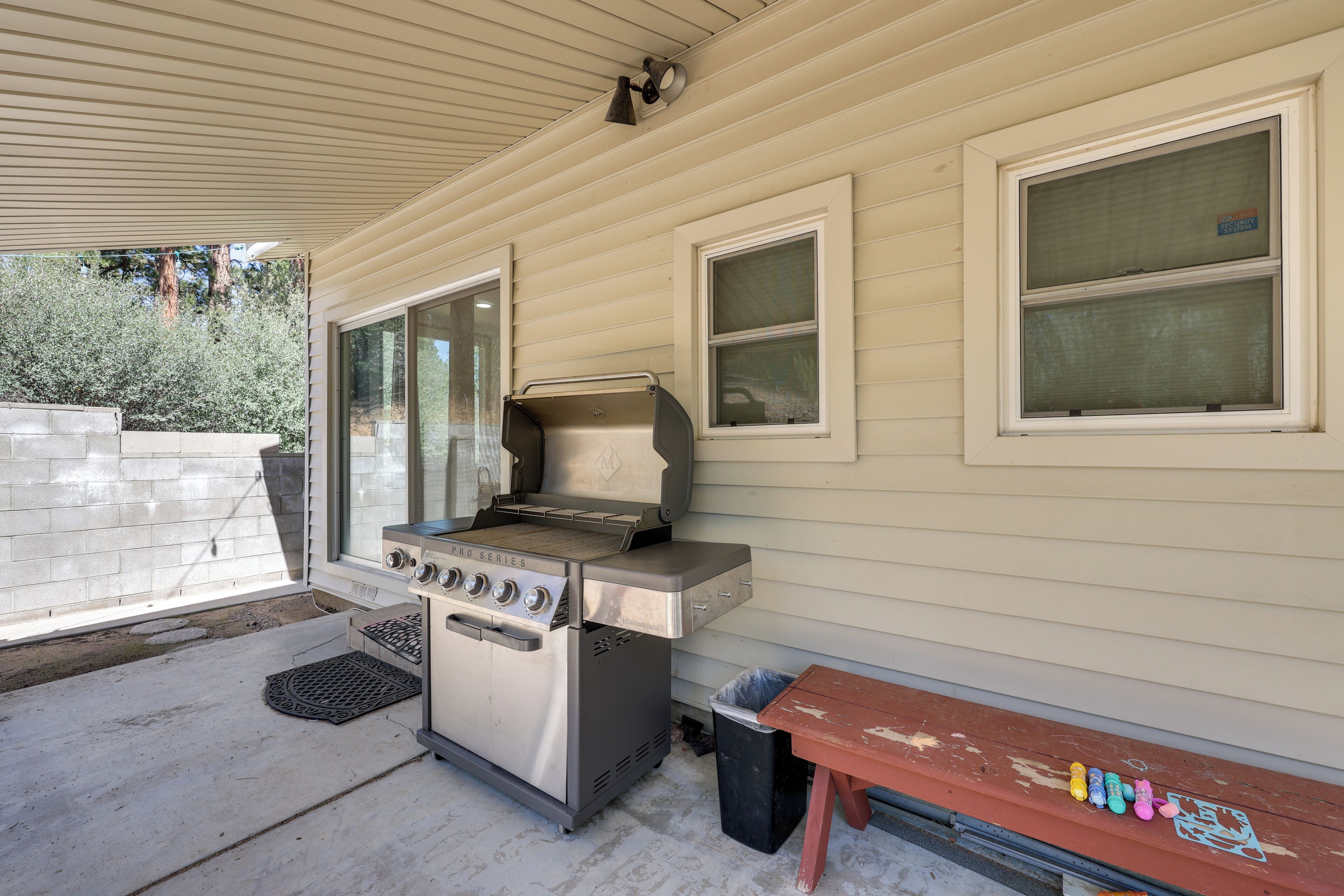 A covered patio with a stainless steel grill, a wooden bench, and a trash can.