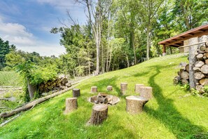 Ein rustikales Outdoor-Ambiente mit einem Feuerkorb, umgeben von Baumstümpfen, einer Holzhütte und einem grasbewachsenen Hügel mit Bäumen im Hintergrund.