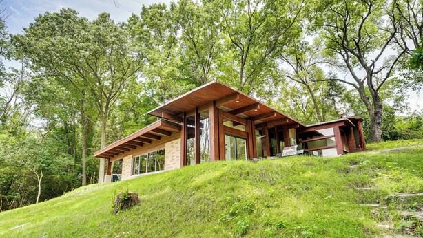 Een modern huis met een schuin dak, grote ramen en een houten gevel, gelegen op een groene heuvel omringd door bomen.