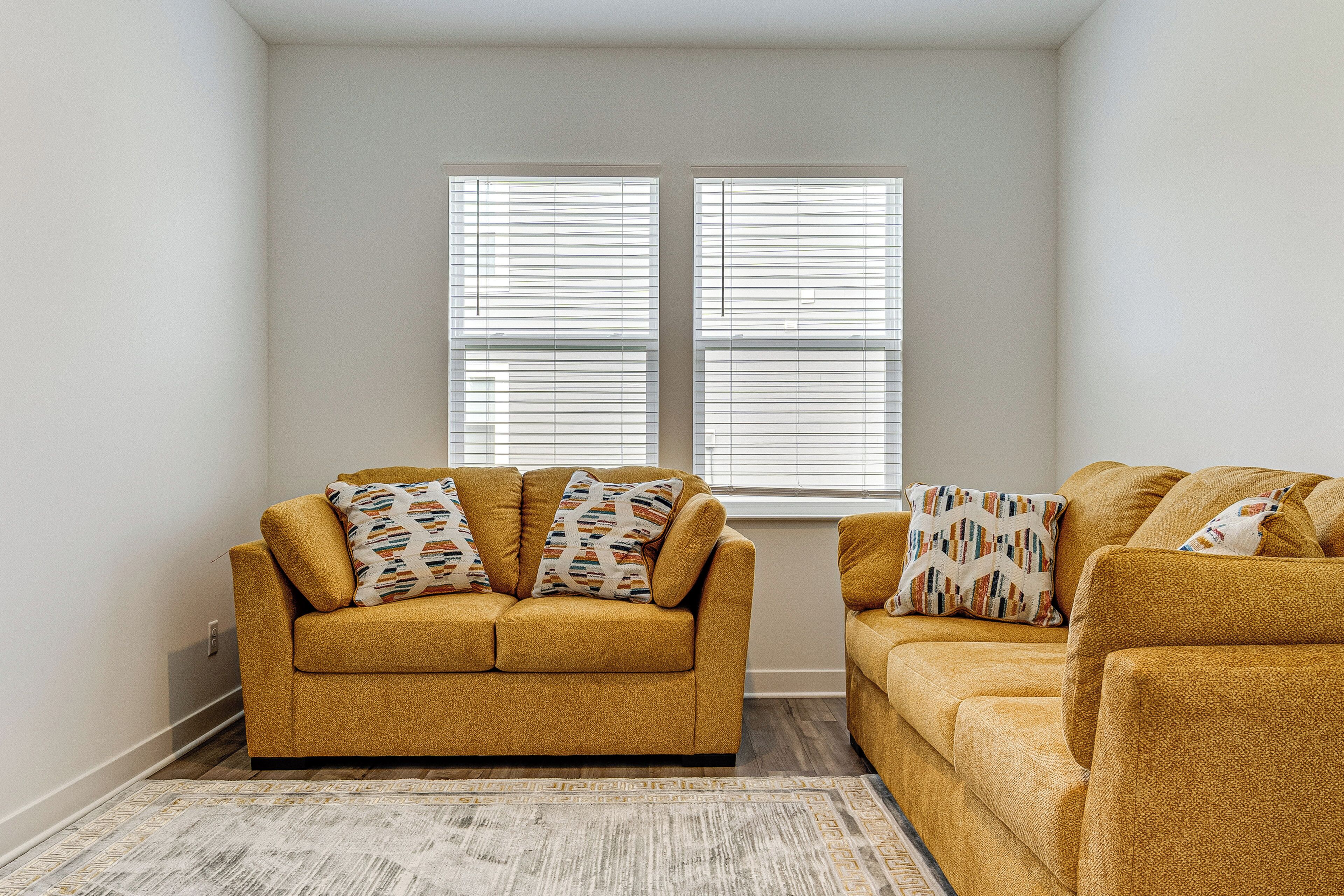 A corner room with two mustard-colored sofas, patterned throw pillows, and a light-colored rug.
