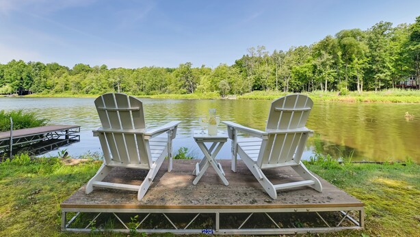 Deux fauteuils Adirondack sur une terrasse en bois, au bord d’un lac, avec une forêt en arrière-plan.